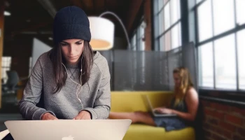 dziewczyna siedząca przed laptopem ze słuchawkami na uchu i w czarnej czapce. Z tyłu druga dziewczyna pracująca na laptopie, siedzi na żółtej kanapie.