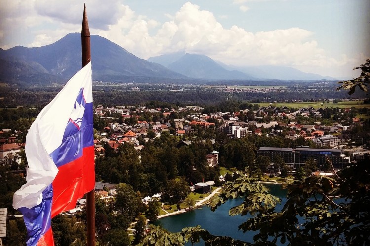 Zdjęcie przedstawia panoramę jednego ze słoweńskich miast. Na pierwszym planie widać trzepoczącą na wietrze flagę Słowenii. Z kolei w tle widać góry.