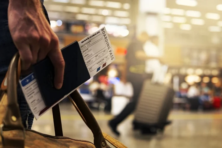 ręka trzymająca bilet lotniczy na lotnisku. Dalej osoba idąca z walizką.