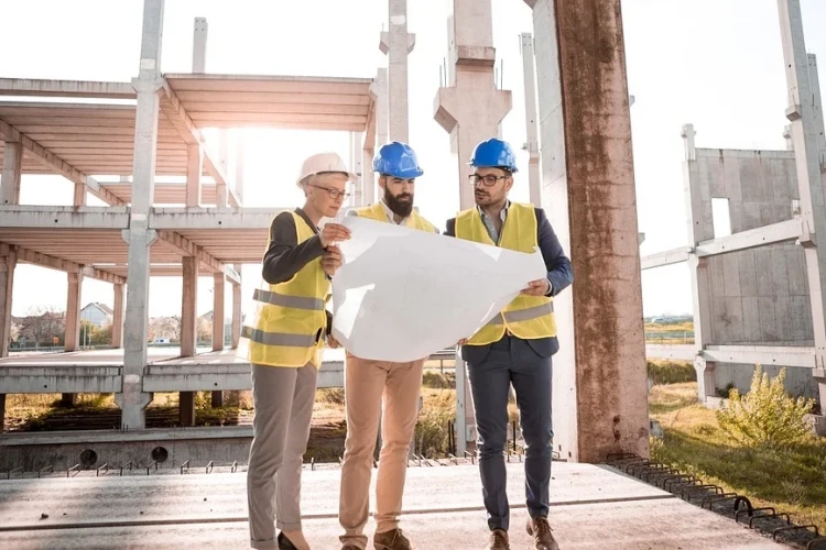 Architekt w kasku wraz z kierownikiem budowy oraz członkiem zespołu projektowego czytający plan budynku na tle powstającego kompleksu przemysłowego